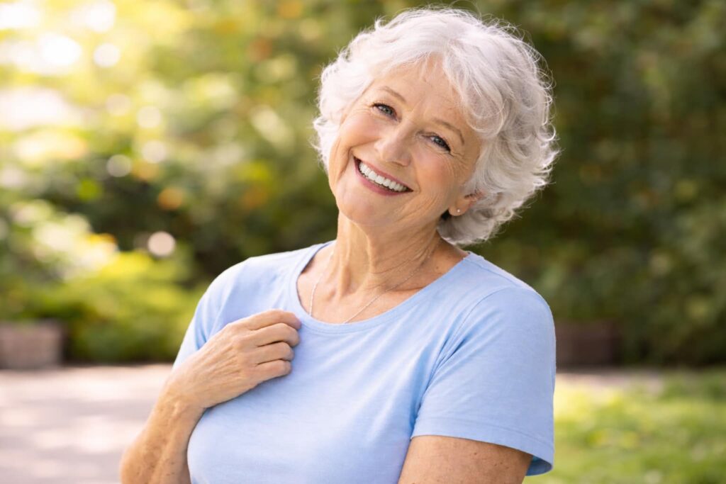 Glücklich lächelnde Seniorin in heller Kleidung steht entspannt im Garten und blickt freundlich in die Kamera. Ein Symbol für Lebensqualität, Sicherheit und Wohlbefinden durch Physiotherapie im Alter.