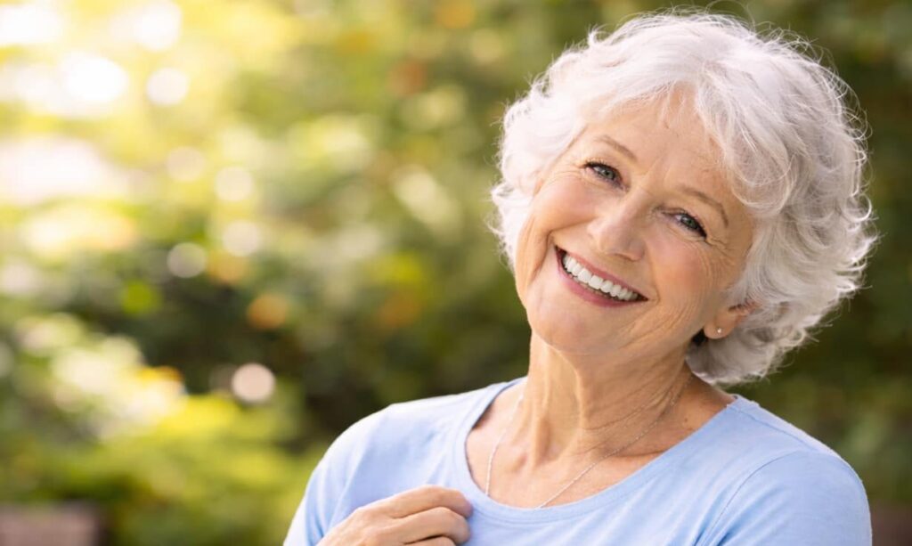 Glücklich lächelnde Seniorin in heller Kleidung steht entspannt im Garten und blickt freundlich in die Kamera. Ein Symbol für Lebensqualität, Sicherheit und Wohlbefinden durch Physiotherapie im Alter.